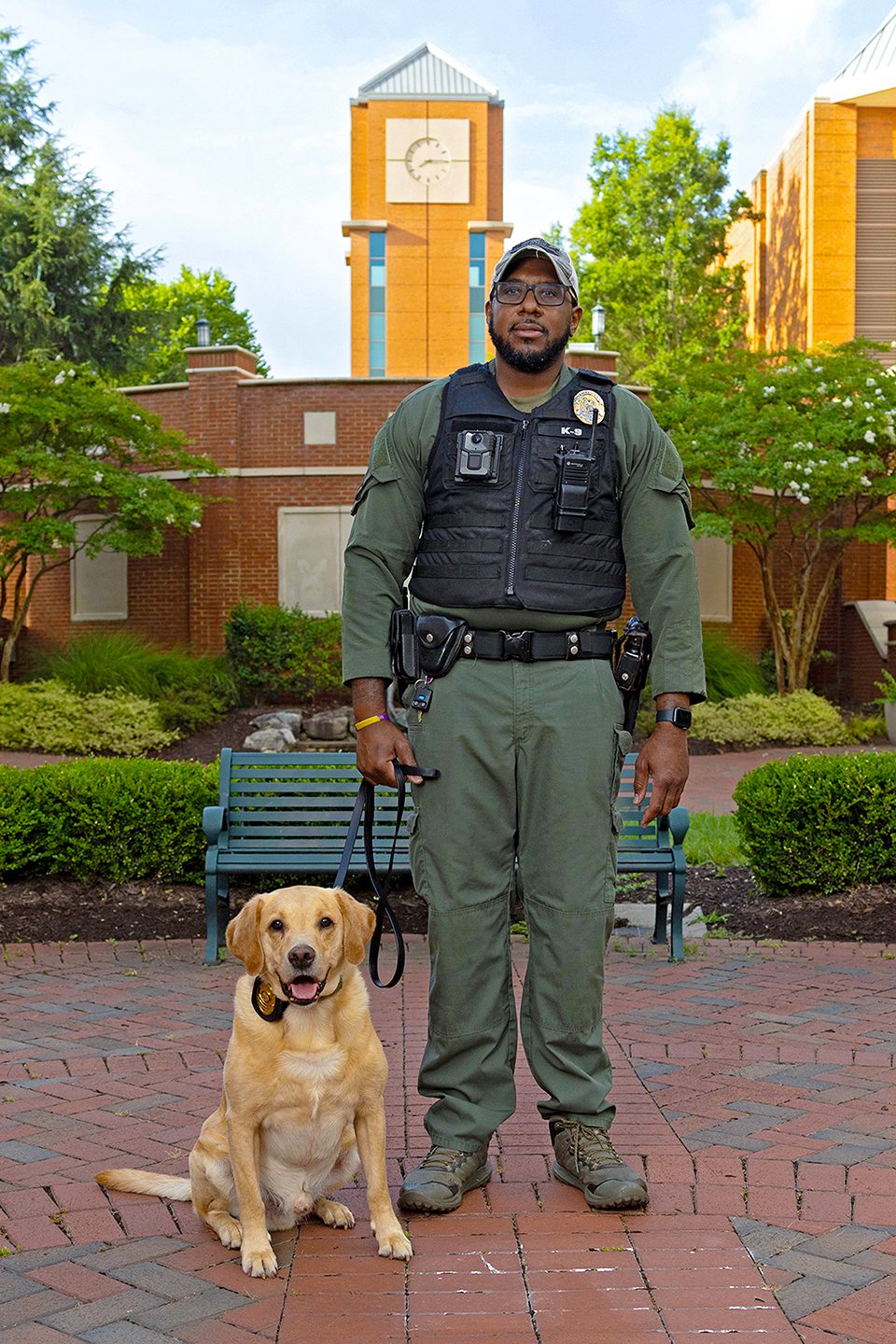 PPS_K9s_25003_5231_kpl A police officer stands with a yellow Labrador Retriever sitting at his side in front of a campus clock tower.