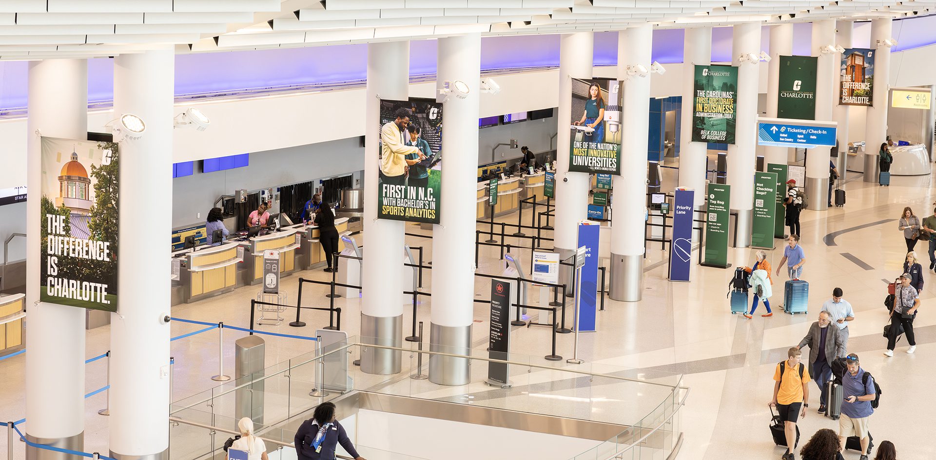 UNC Charlotte promotional banners line tall pillars above the airport’s check-in counters. Messaging includes “The Difference is Charlotte,” “First in N.C. with Bachelor’s in Sports Analytics,” and “One of the Nation’s Most Innovative Universities.