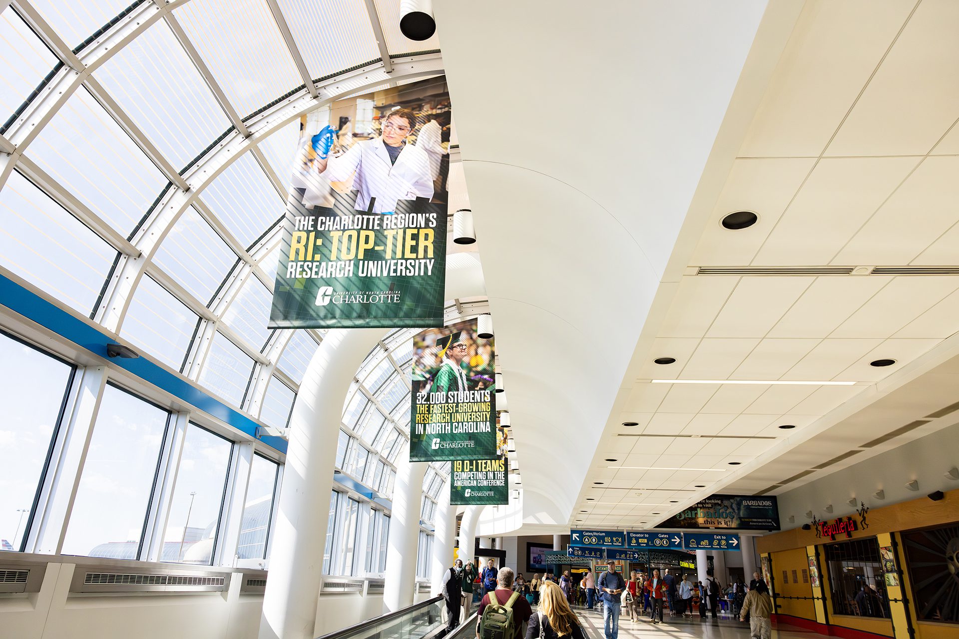 Passengers walk beneath a series of UNC Charlotte banners in a bright, window-lined airport concourse. The signs highlight Charlotte’s R1 research status, enrollment growth to 32,000 students, and NCAA Division I athletics. Each banner features engaging images of students, faculty, and campus life.