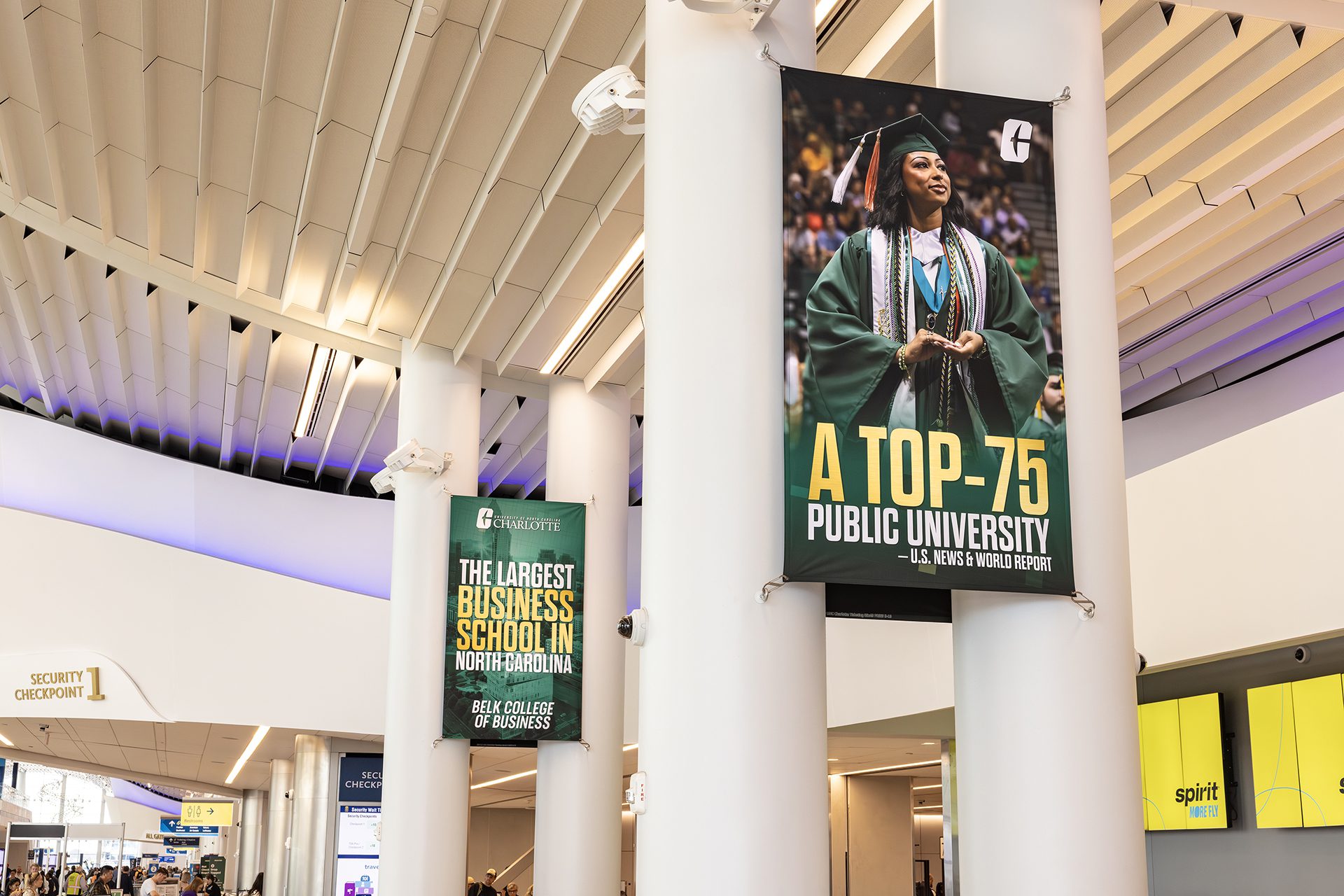 Large UNC Charlotte banners hang inside Charlotte Douglas International Airport. One features a graduate in green regalia with the headline “A Top-75 Public University – U.S. News & World Report.” Another banner highlights “The Largest Business School in North Carolina – Belk College of Business.” Travelers pass through Security Checkpoint 1 nearby.