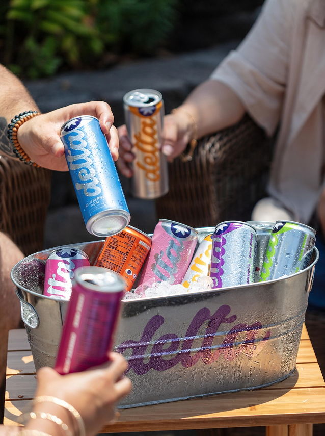 Drink-Delta1 People reach into a metal bucket filled with ice and colorful Delta seltzer cans during an outdoor gathering, each holding a can of different flavor and dosage.