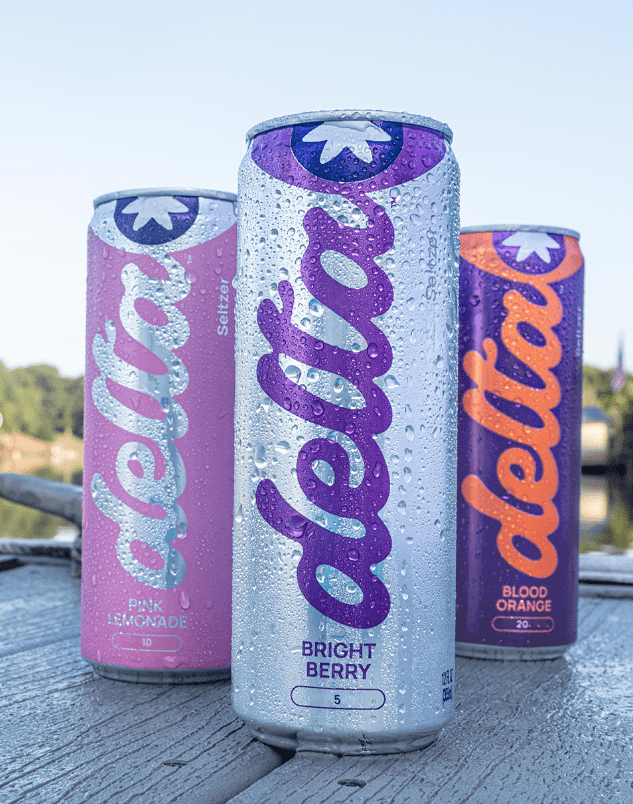 Drink-Delta2 Three condensation-covered Delta seltzer cans in Bright Berry, Pink Lemonade, and Blood Orange flavors stand upright on a wooden surface near the water.