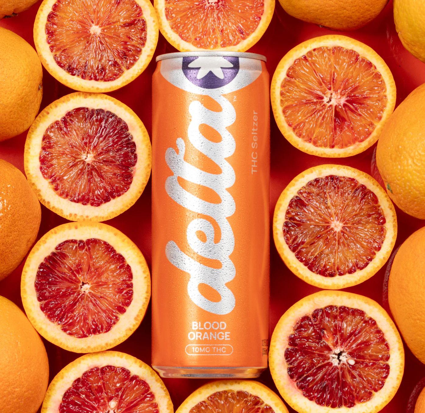 Drink-Delta5 A can of Delta THC Seltzer in Blood Orange flavor sits upright on a vibrant red surface, surrounded by halved and whole blood oranges, emphasizing its citrusy, refreshing appeal.