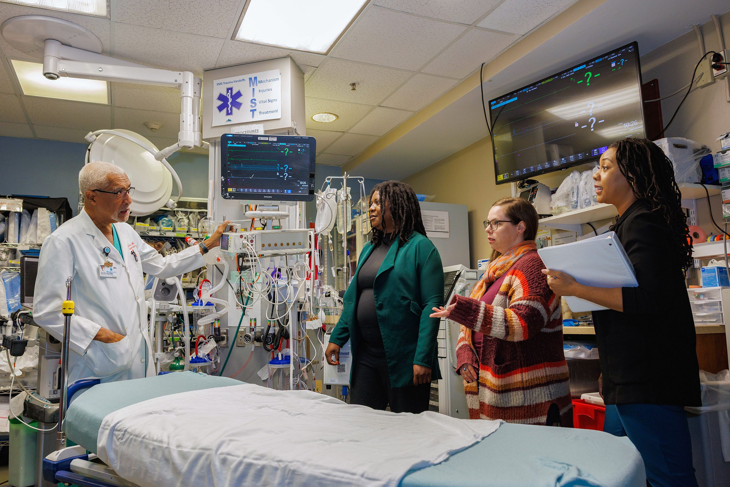A hospital professional in a white coat leads a group discussion around medical equipment in a clinical training room, with three other people observing attentively.
