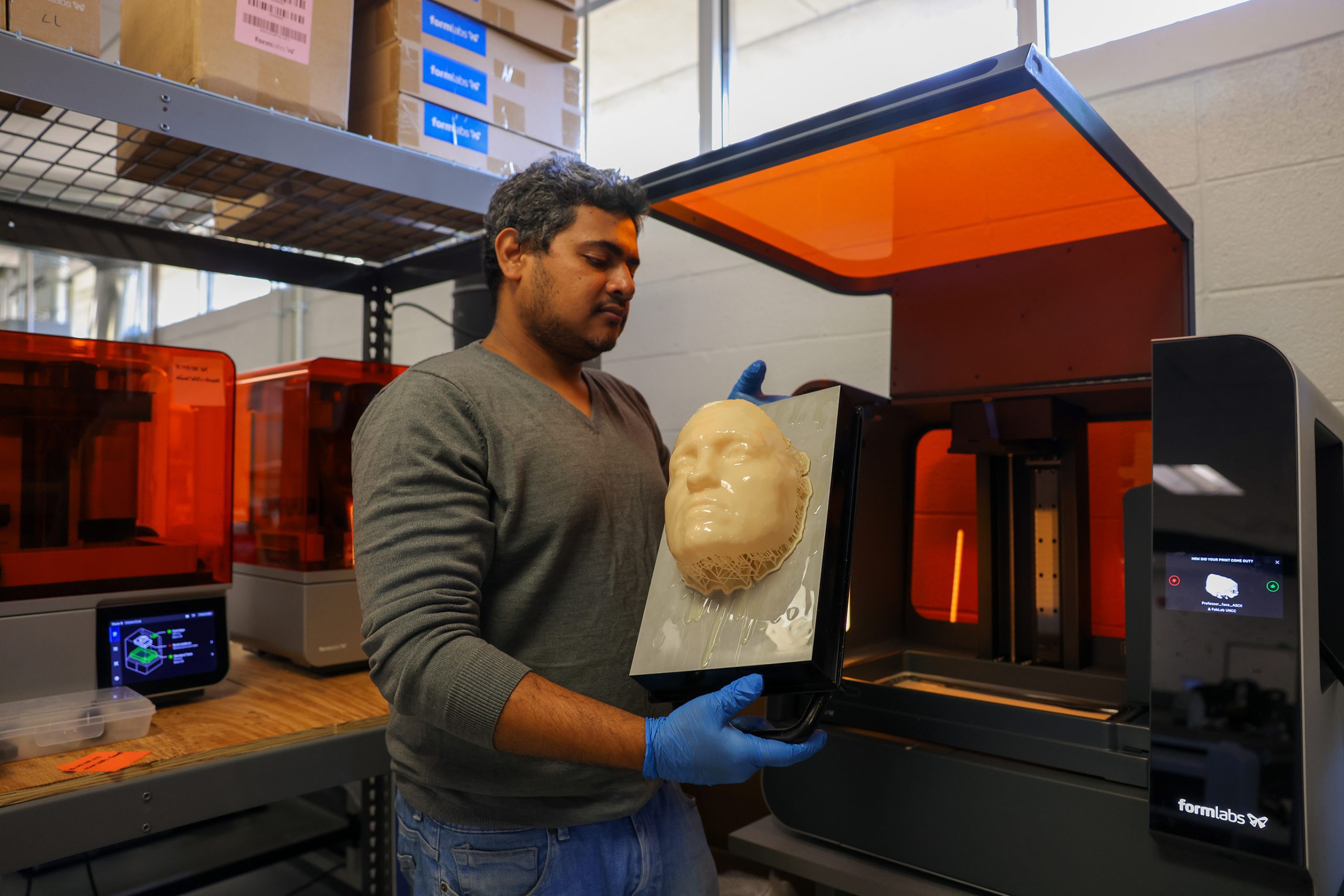 A researcher wearing gloves holds a freshly printed 3D face model from a large Formlabs 3D printer in a university lab setting.