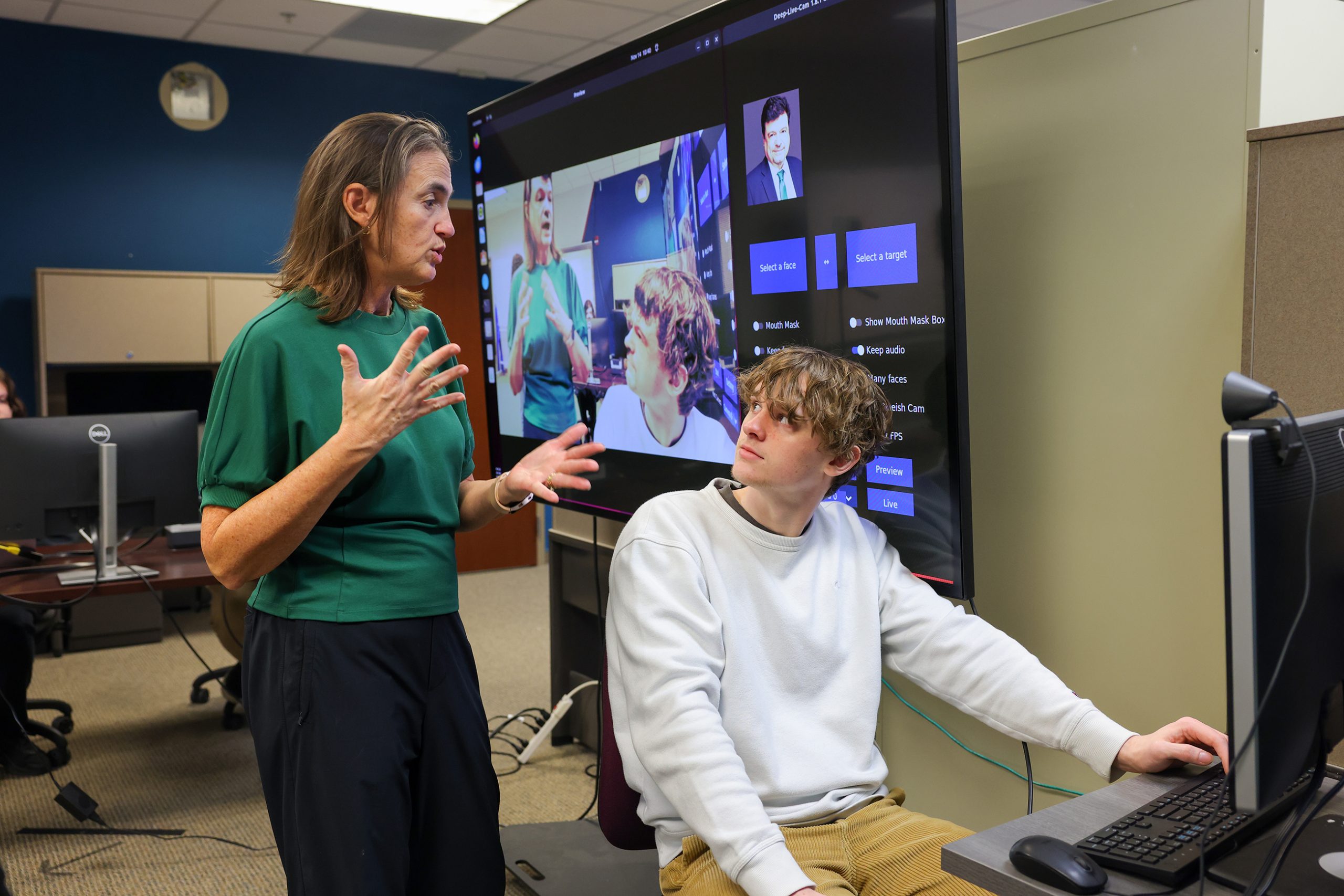 A UNC Charlotte instructor gestures while speaking to a student at a computer, with a large monitor behind them displaying face manipulation software and deepfake visuals.