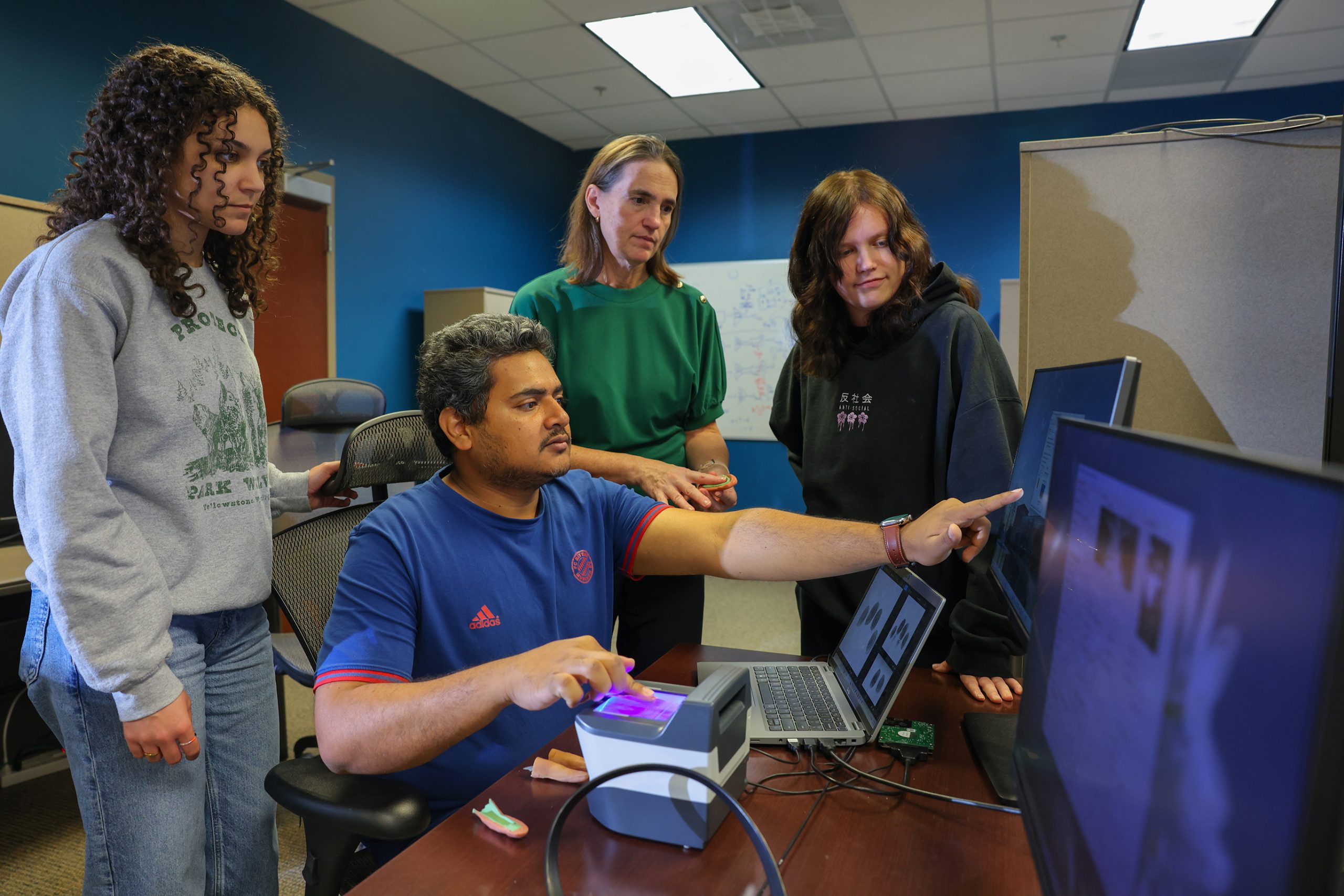 A group of students and a researcher gather around a desk in a computer lab as one student demonstrates biometric scanning on a laptop connected to various devices.