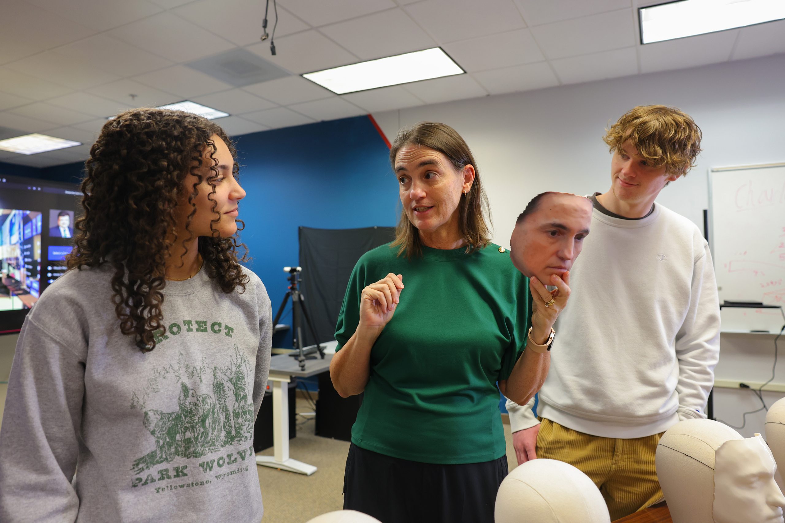 A professor holds up a realistic 3D-printed face mask while talking to two students in a lab. Other mannequin heads with facial models are placed on the table nearby.
