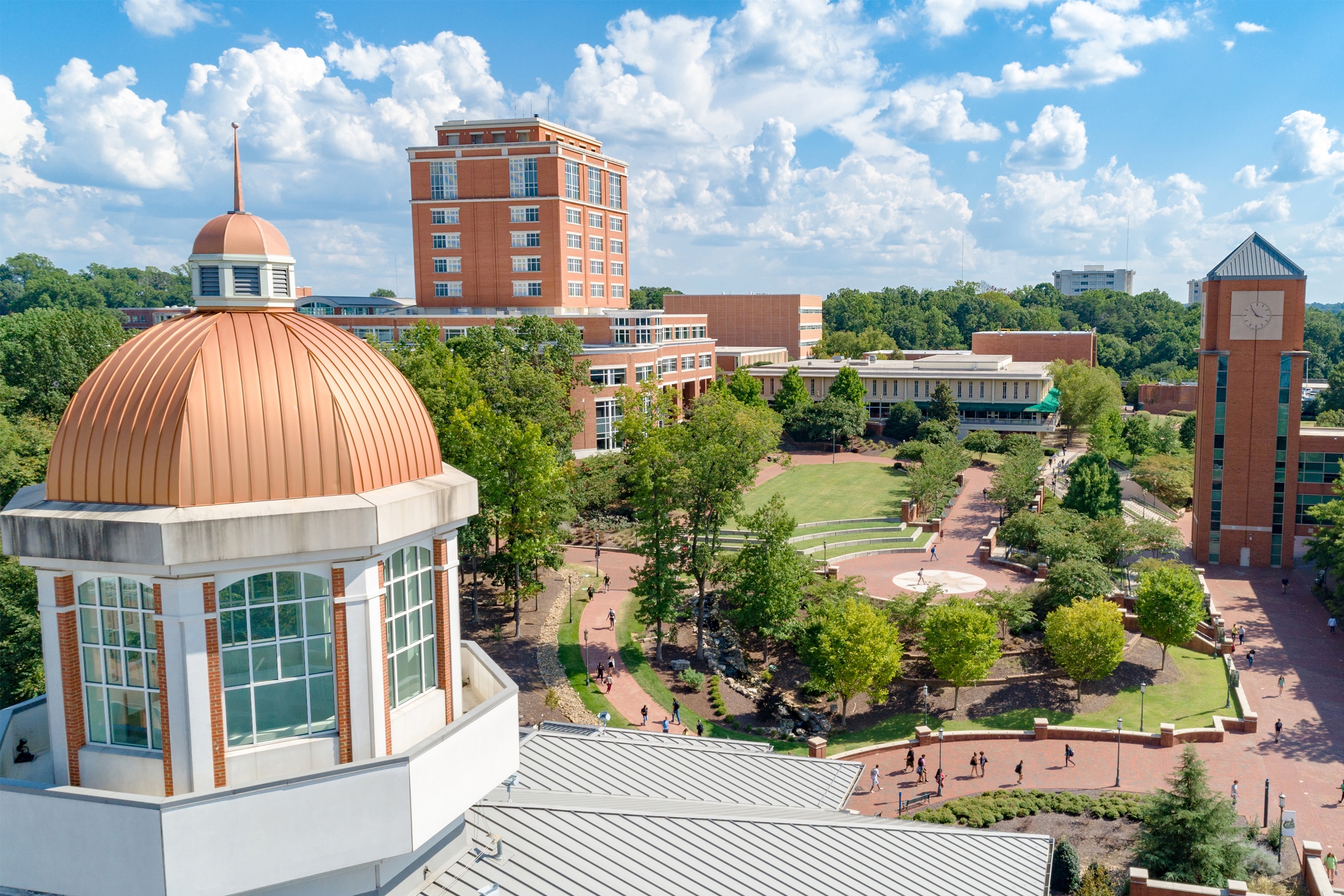 Aerial_Atkins_Clocktower_Cupola Elevated view of UNC Charlotte campus with a prominent domed building, surrounding brick structures, tree-lined paths, and students walking across a landscaped quad.