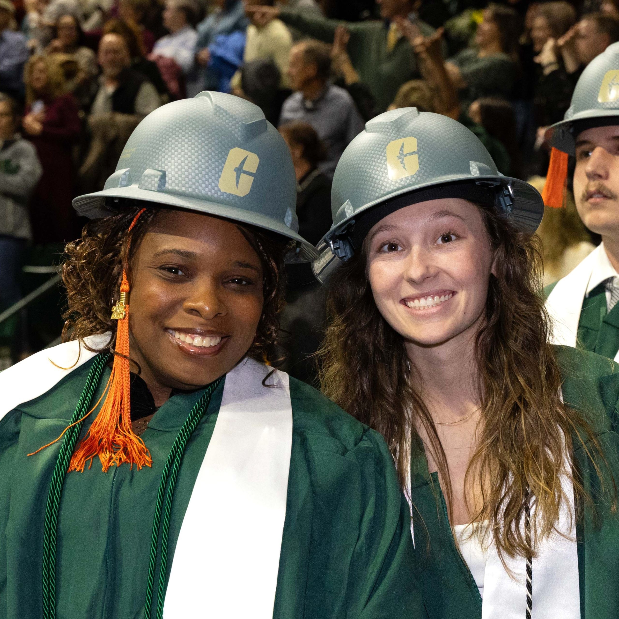 Two construction graduates smile while wearing green hard hats and graduation regalia.
