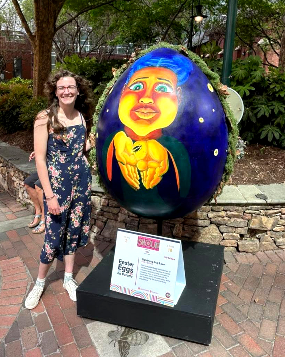Charlotte_SHOUT!_Easter Egg_ Lightning Bug Love_Emily-Little Smiling woman in a floral jumpsuit stands beside a large painted Easter egg sculpture during the Easter Eggs on Parade public art exhibit.