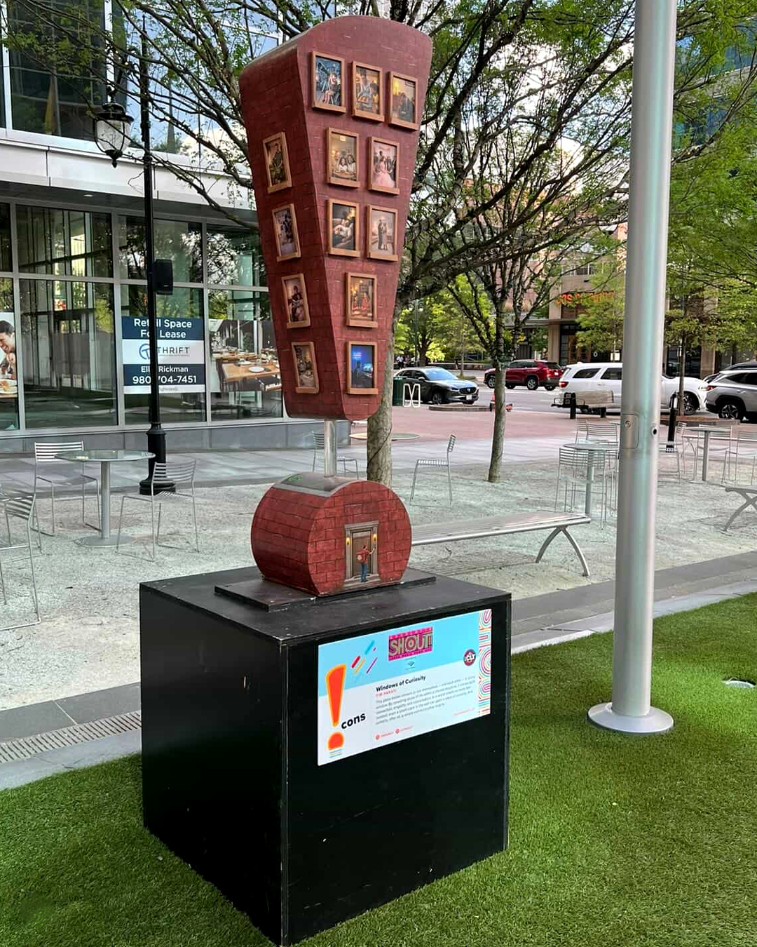 Charlotte_SHOUT!_Windows of Curiosity_Tim-Parati Tall red public art sculpture covered with framed miniature artworks stands on display in an outdoor plaza with trees and seating nearby.