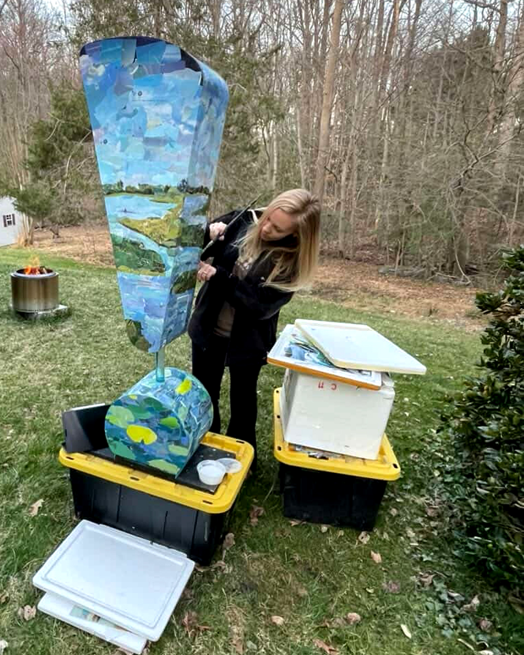 Charlotte_SHOUT!_!con River of Paper_Constance-Sartor Woman adjusts a tall painted outdoor sculpture featuring blue sky and landscape imagery while standing in a grassy backyard beside storage bins and art supplies.