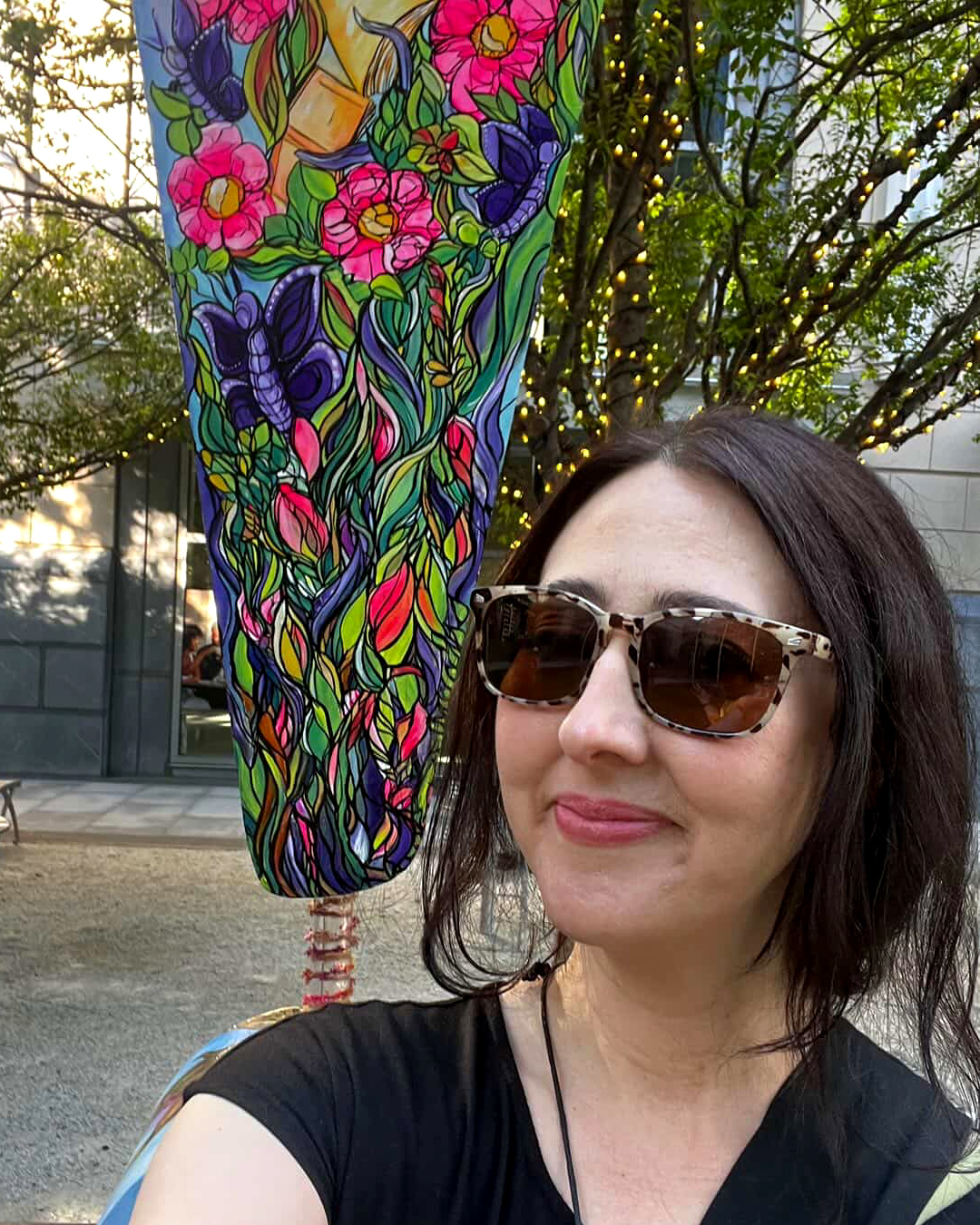 Charlotte_SHOUT!_!con_Blooming Magic_Maria-Napier Woman wearing sunglasses poses beside a colorful floral-painted public art sculpture decorated with bright flowers and flowing green vines.