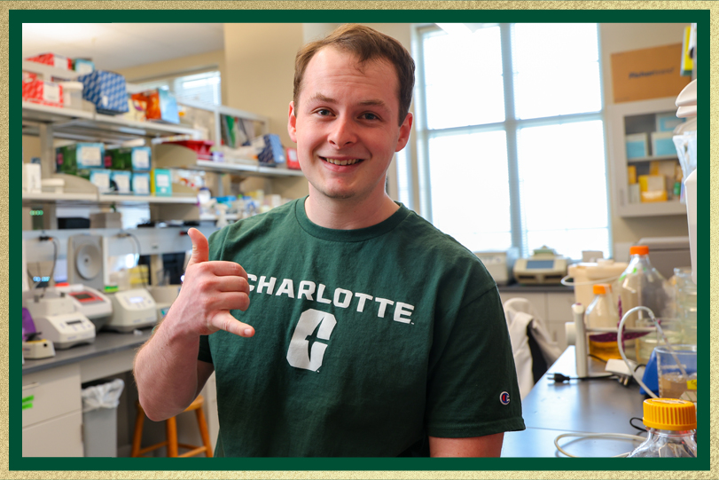 Smiling student in a green Charlotte T-shirt makes a hand gesture while standing in a laboratory with equipment and shelves in the background.