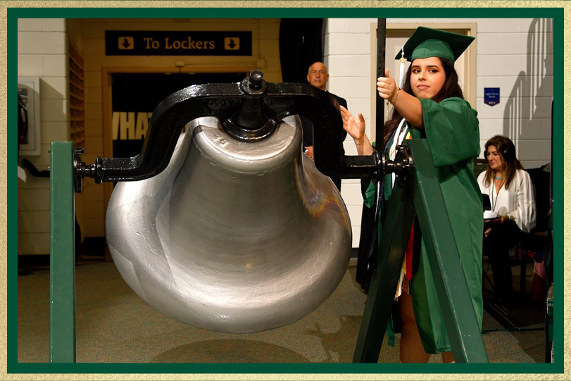 Graduate in a green cap and gown rings a large ceremonial bell indoors during a commencement event, with people and signage visible in the background.