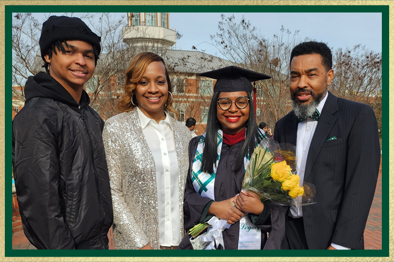 Graduate in cap and gown holds a bouquet of flowers while posing with three family members on a brick campus walkway, with a domed building in the background.