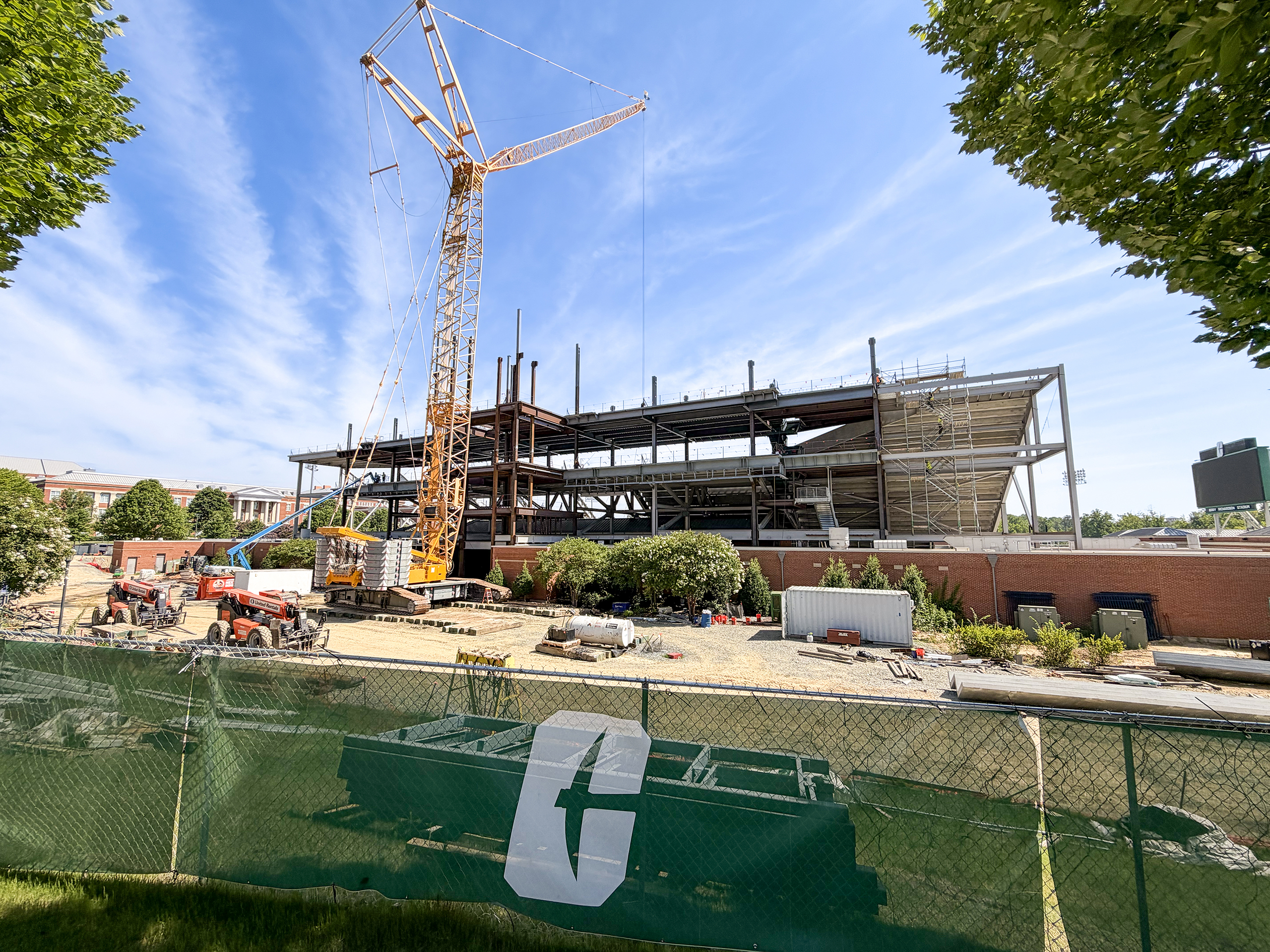Behind Fence Stadium Exterior view of the Jerry Richardson Stadium expansion under construction, showing a large crane, exposed steel framework, and active construction site behind a green fence with Charlotte branding beneath a bright blue sky.