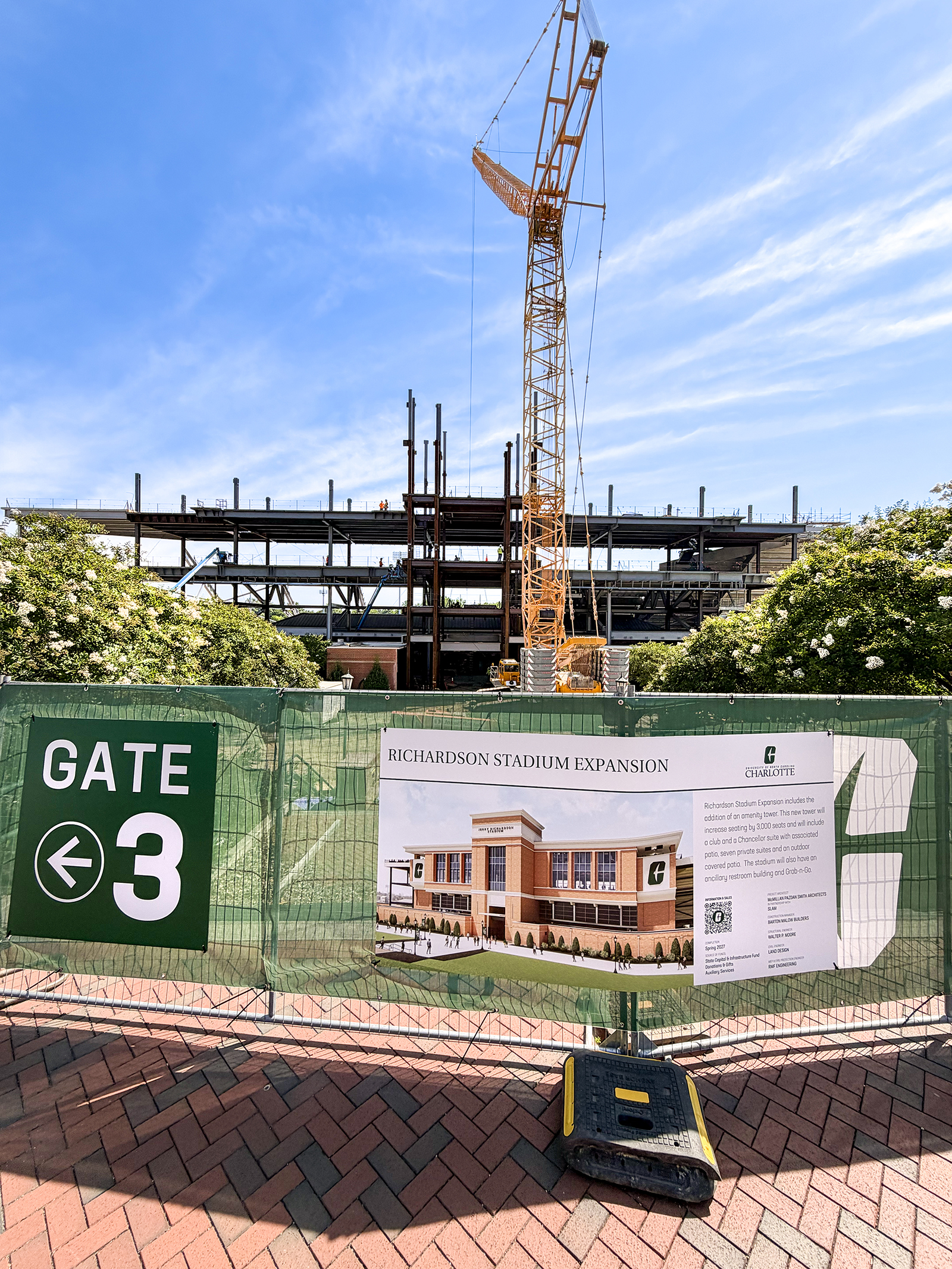 Entrance Sign Stadium View of the Jerry Richardson Stadium expansion construction site from outside Gate 3, with a towering crane, exposed steel framework, and a construction fence displaying a project rendering and information about the stadium expansion.