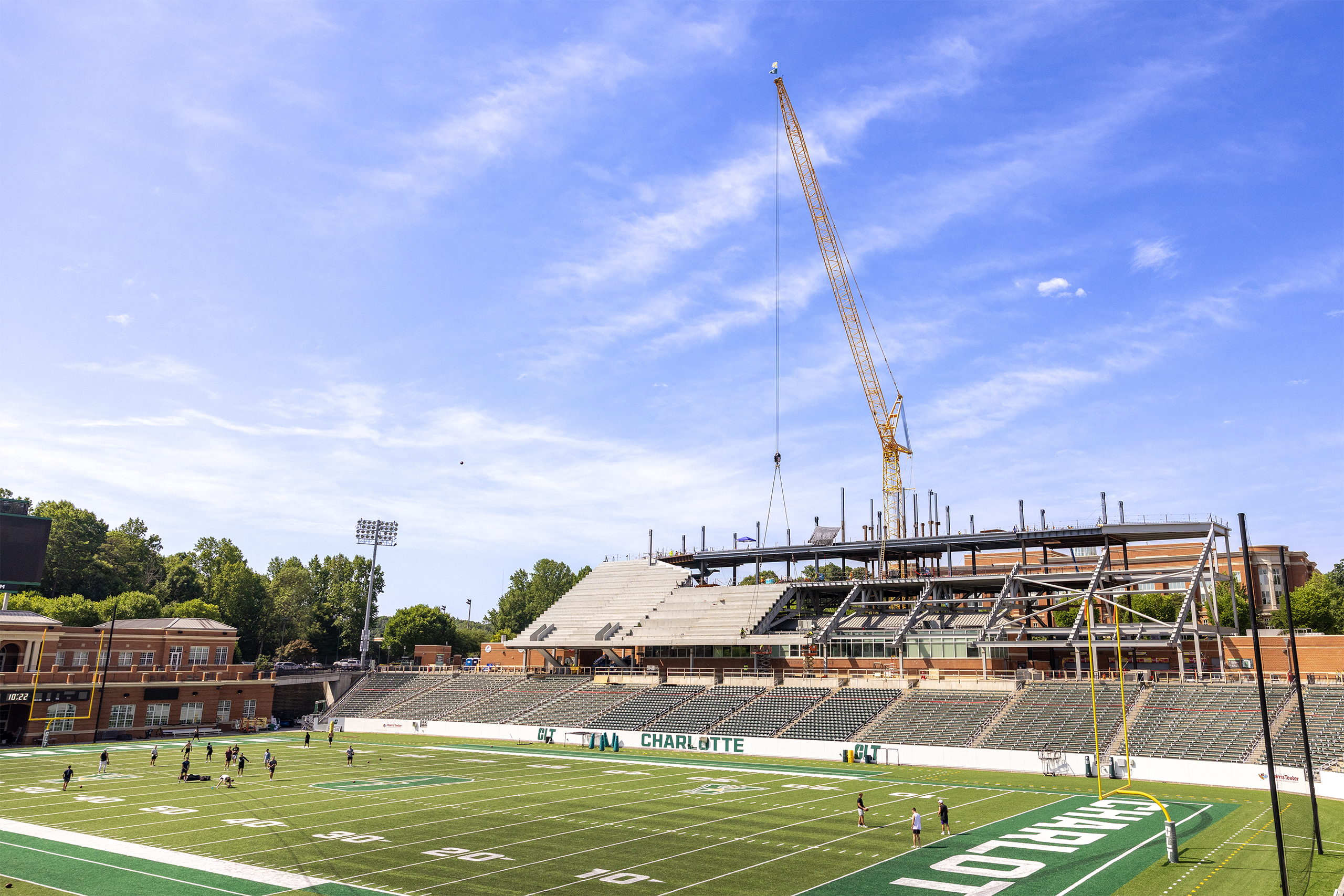 Jerry Richardson Stadium undergoes expansion construction as a large crane towers above newly installed steel beams and concrete seating. The football field remains in use below, with a few people on the turf beneath a bright blue sky.