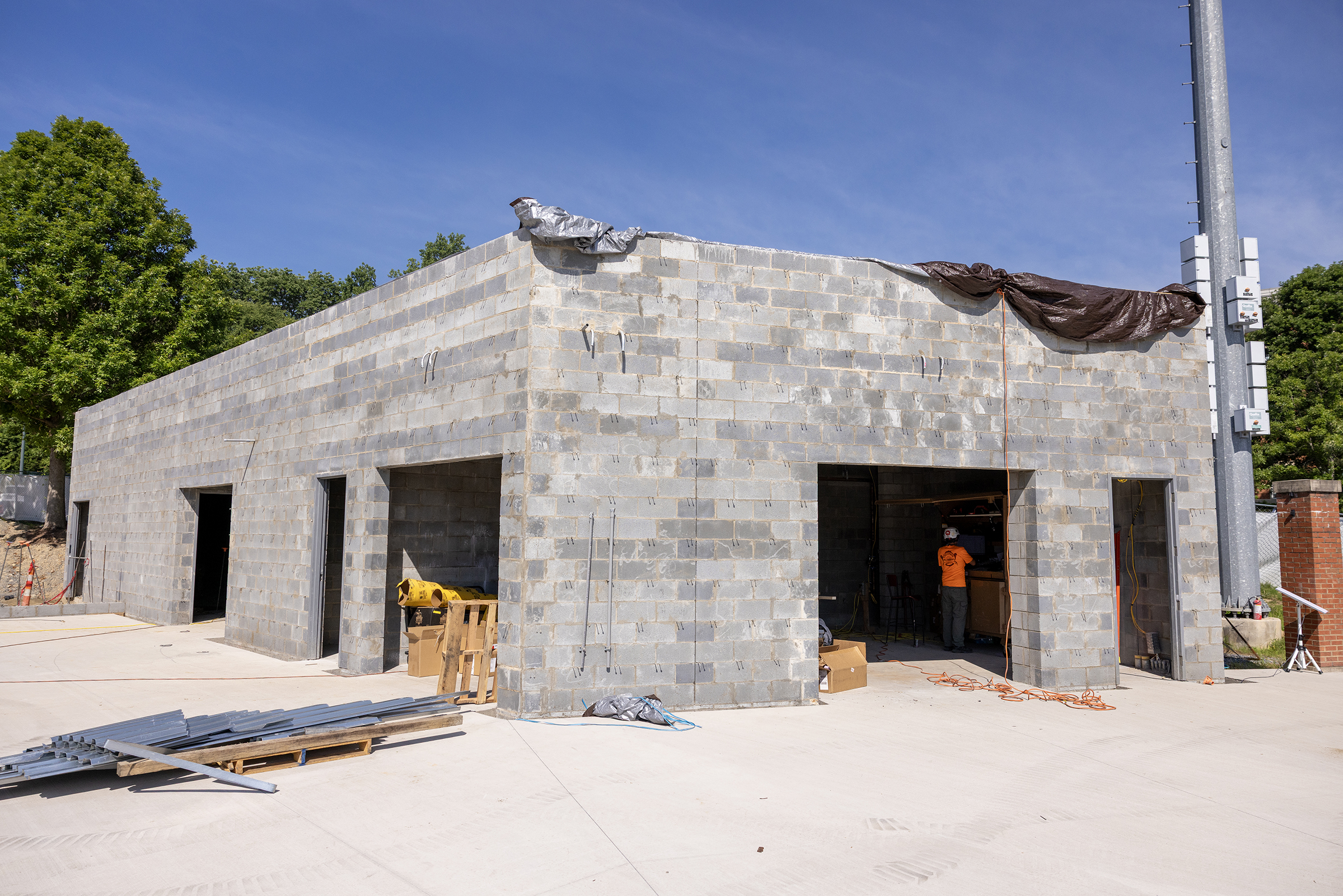 Grab and Go Stadium Concrete block utility building under construction near Jerry Richardson Stadium, with open doorways, construction materials on the ground, and a worker visible inside. A protective tarp covers part of the unfinished roof beneath a bright blue sky.
