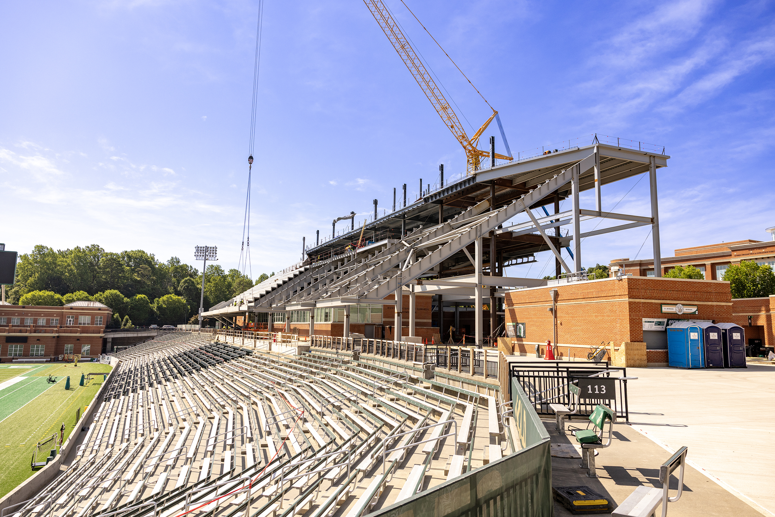 Near Side Stadium Jerry Richardson Stadium's west grandstand under construction, with exposed steel framing, newly installed seating, and a towering crane above the expansion. Existing stadium seating and the football field remain visible in the foreground beneath a clear blue sky.