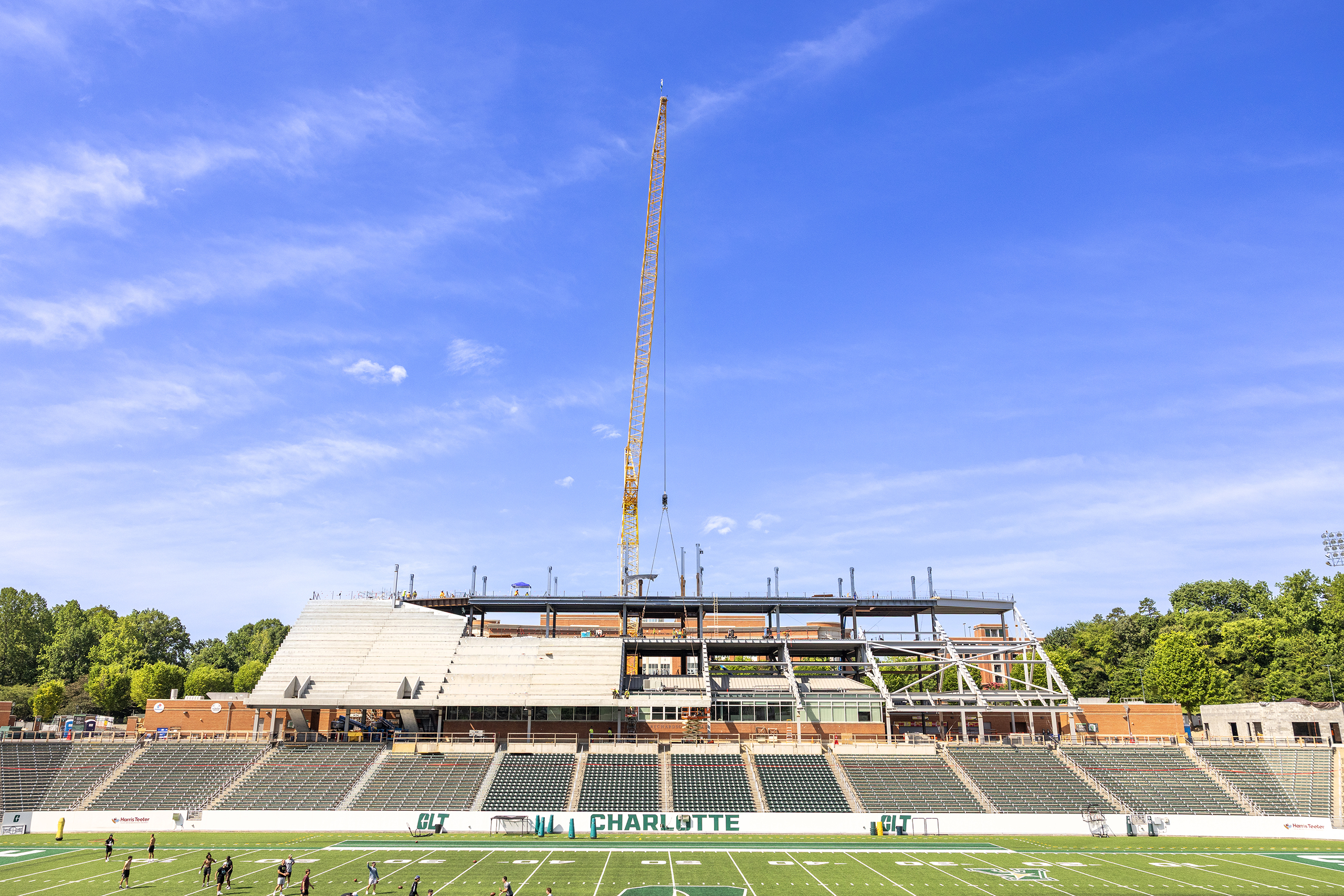 Straight Across Stadium Front view of Jerry Richardson Stadium during construction, showing a large crane above the expanding west grandstand with exposed steel framework and partially completed concrete seating. The football field and existing stands remain in use below under a bright blue sky.