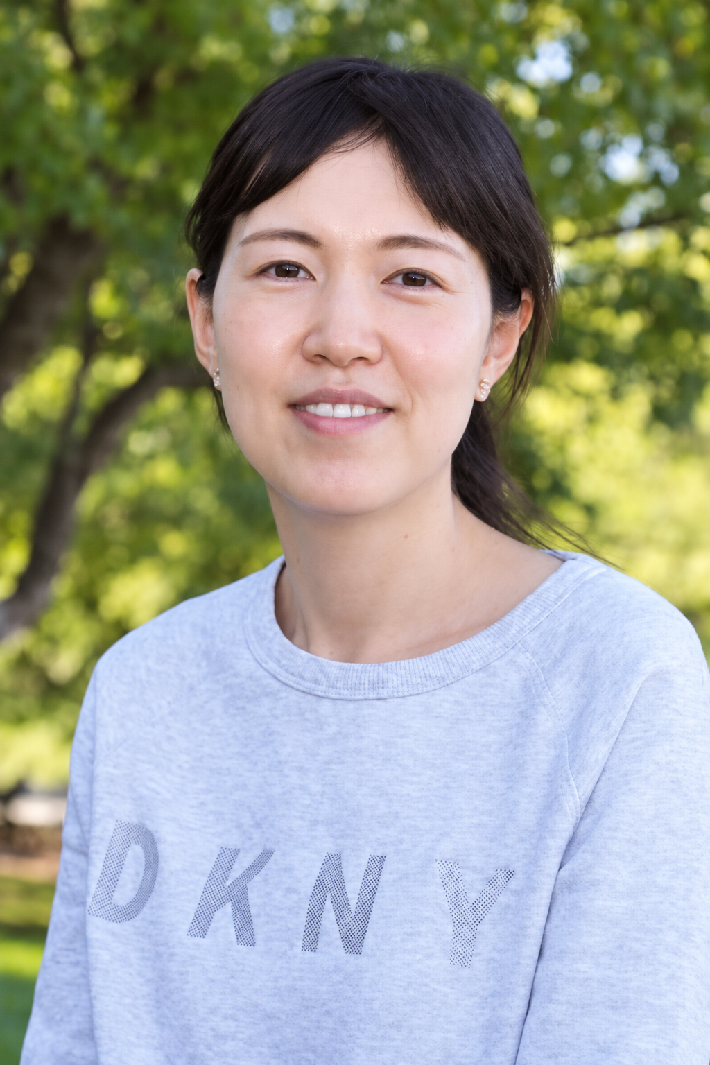 2026 Truist Report - Wenwen Dou Headshot Outdoor portrait of a smiling woman with dark hair wearing a light gray sweatshirt, with leafy green trees softly blurred behind her.