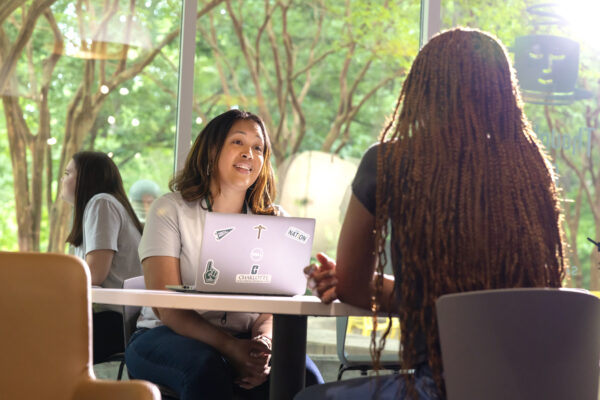 Two individuals engage in conversation at a table with a laptop, set against a backdrop of lush greenery and natural light.