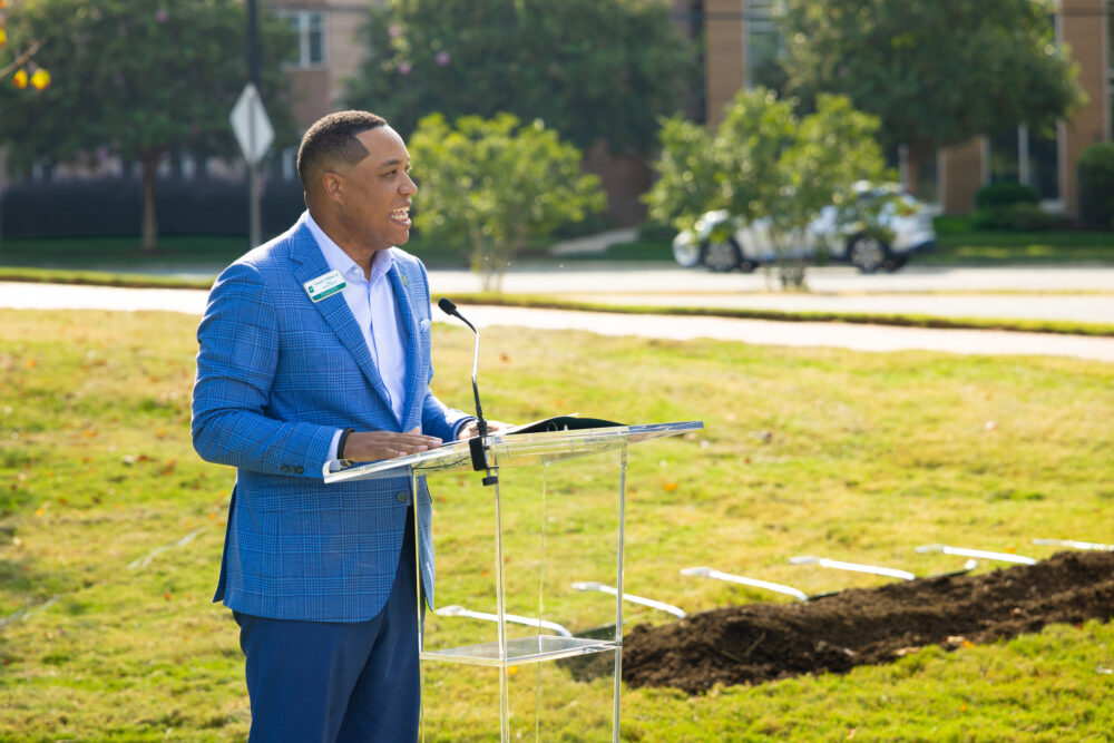 A person in a blue plaid suit stands at a clear podium outdoors, speaking into a microphone on a grassy field with construction debris nearby.