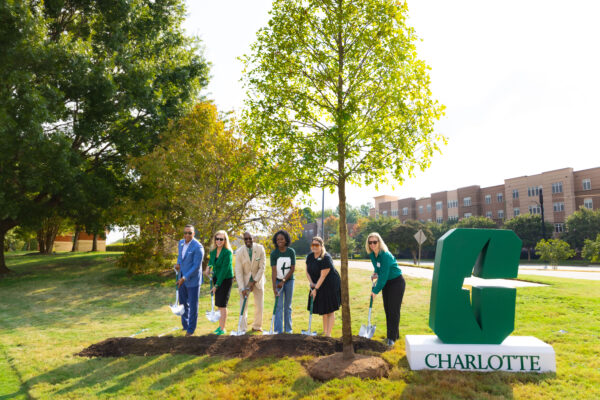 Six individuals participate in a tree planting ceremony beside a large green “C” sign that reads “Charlotte,” with a newly planted tree and shovels in the foreground.