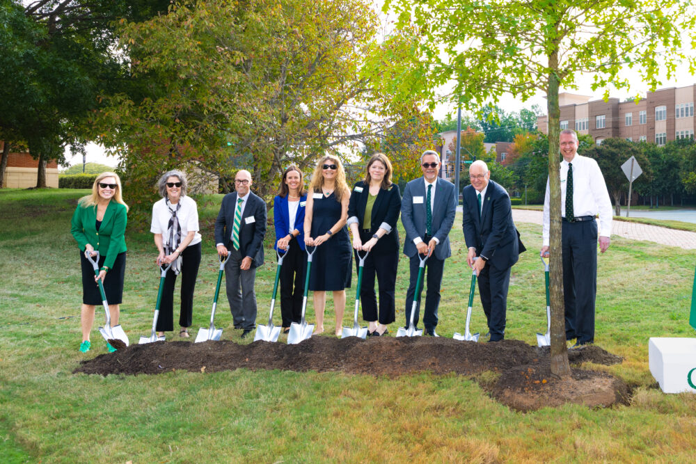 A group of eight individuals stands outdoors with shovels, participating in a tree planting ceremony on a grassy area.
