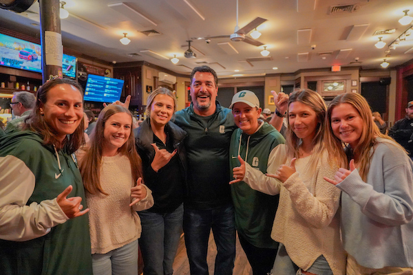 A group of people in green apparel pose together at a bar, smiling and making hand gestures.