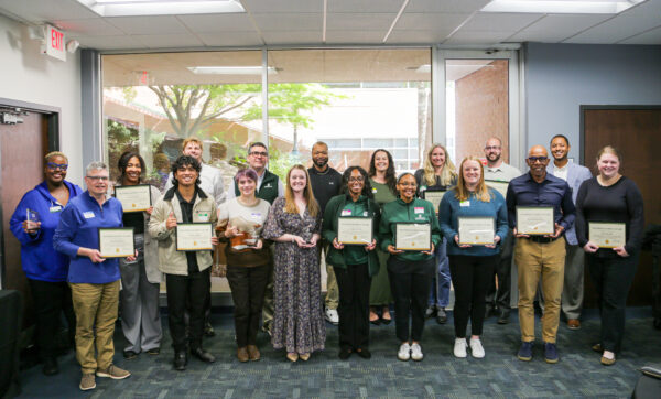 A group of individuals in a room hold certificates and trophies, posing for a photo with smiles and celebratory expressions.