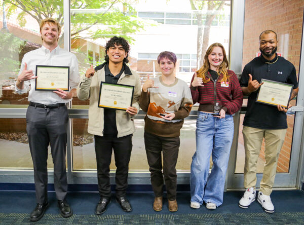 Five individuals stand together indoors, each holding a framed certificate and making a thumbs-up gesture.
