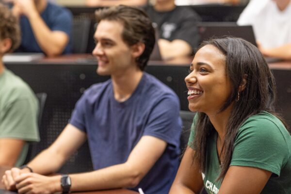 Two students smile and engage in a lecture hall, with one looking toward the speaker and the other smiling broadly.