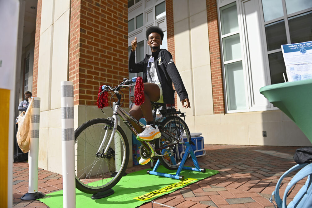 A person smiles while sitting on a bicycle mounted on a stand outdoors, gesturing upward with one hand.