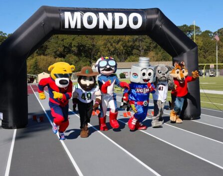 Multiple mascots line up at a track start line under a “MONDO” arch.