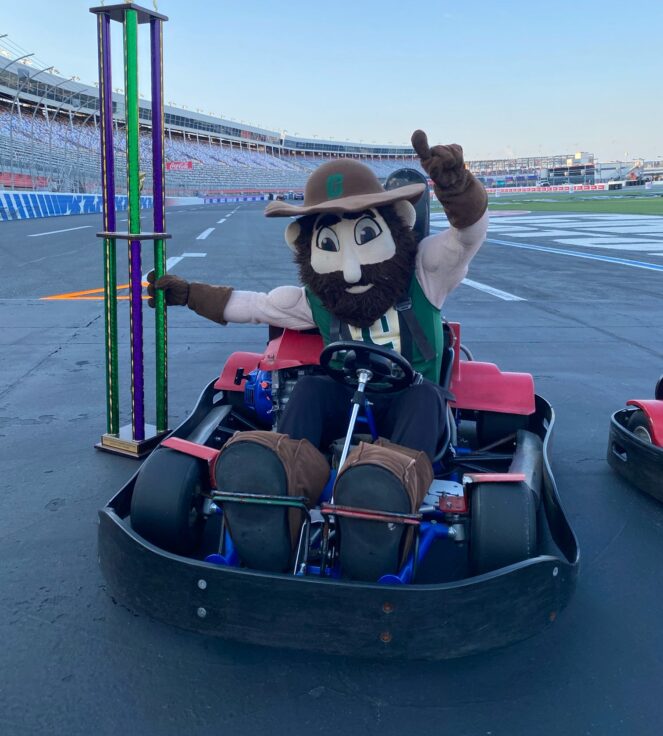 Norm the Niner in a go-kart holding a tall trophy on a racetrack.
