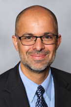 2026 Truist Report - Scott Tonidandel Headshot Professional headshot of a smiling man wearing glasses, a dark suit, light blue shirt and patterned tie against a gray background.