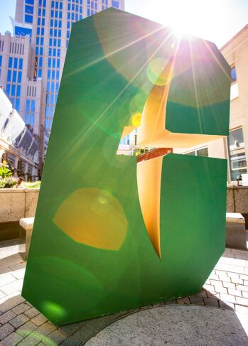All-In-C_NNW Uptown Large green “C” sculpture in an urban plaza with sunlight streaming through its center, creating lens flare, with tall buildings in the background.