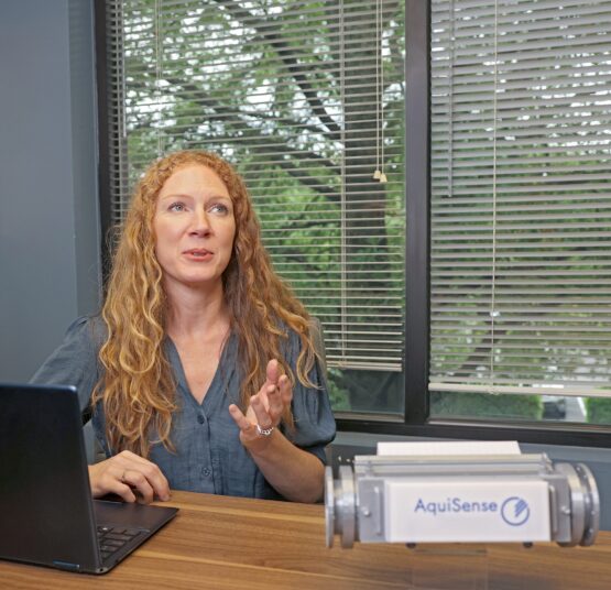 A woman with long curly red hair sits at a wooden desk in front of a laptop, gesturing as she speaks. Beside her is a device labeled “AquiSense,” and a large window with partially open blinds reveals trees outside.