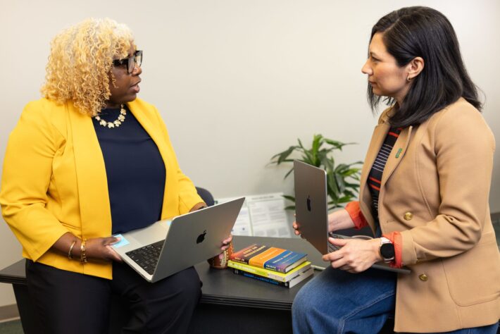 CATO_Carmen-Serrata_Constellation-Mentorship_26026_9939_ah Two women holding laptops sit and talk in an office setting beside a desk with stacked books and a small plant.