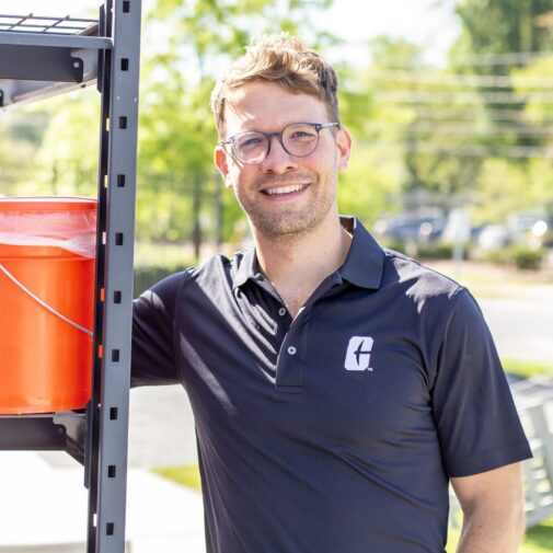 CT-Compost-Kris-Steele Man in UNC Charlotte polo stands next to compost buckets on outdoor shelving.