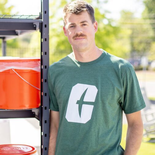 CT-Compost-Eric-Theys Man in a green “C” logo shirt stands beside a shelving unit holding white and orange Crown Town Compost buckets.