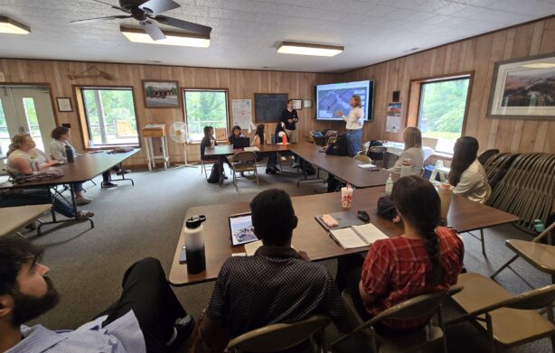 Chimney-Rock_UNC-Charlotte A group of people seated in a wood-paneled meeting room listening to a presentation. Two people at the front of the room are pointing to slides on a screen, while others sit around tables with laptops and notebooks.