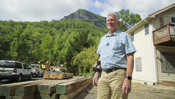 Chimney Rock Revival A man stands outside in front of construction materials and vehicles, with Chimney Rock visible in the background. He wears a short-sleeve patterned shirt and looks toward the camera.