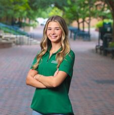 Commencement_Abby_Gordon Student in green university polo stands confidently on a brick pathway lined with trees.