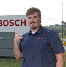 Commencement_Davey_Sides Smiling man in Bosch polo gives shaka sign while standing in front of Bosch corporate sign.