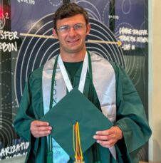 Commencement_Ean_Bass Graduate in cap and gown smiles while holding green cap, standing in front of science-themed mural.